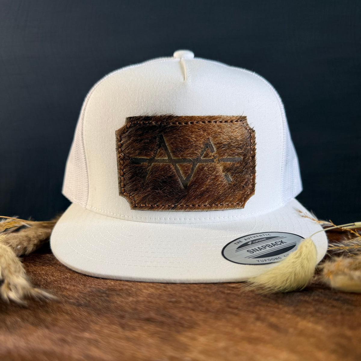 White snapback hat with warm brown scalloped cowhide patch and tonal hand-stitched border, branded with a registered cattle brand