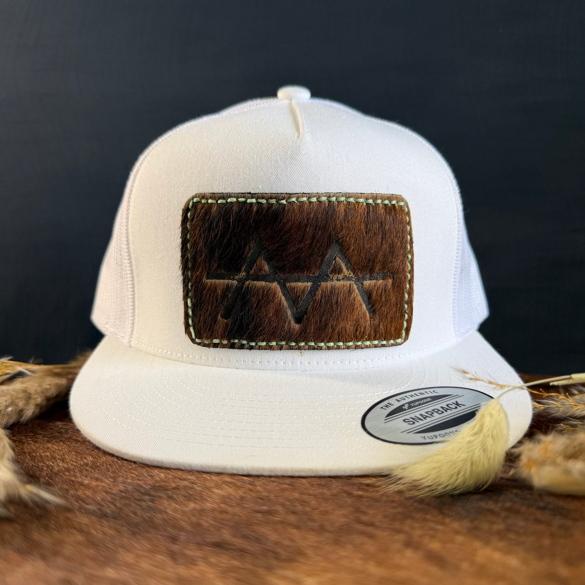 White snapback hat with warm brown cowhide patch and cream hand-stitched border, branded with a registered cattle brand