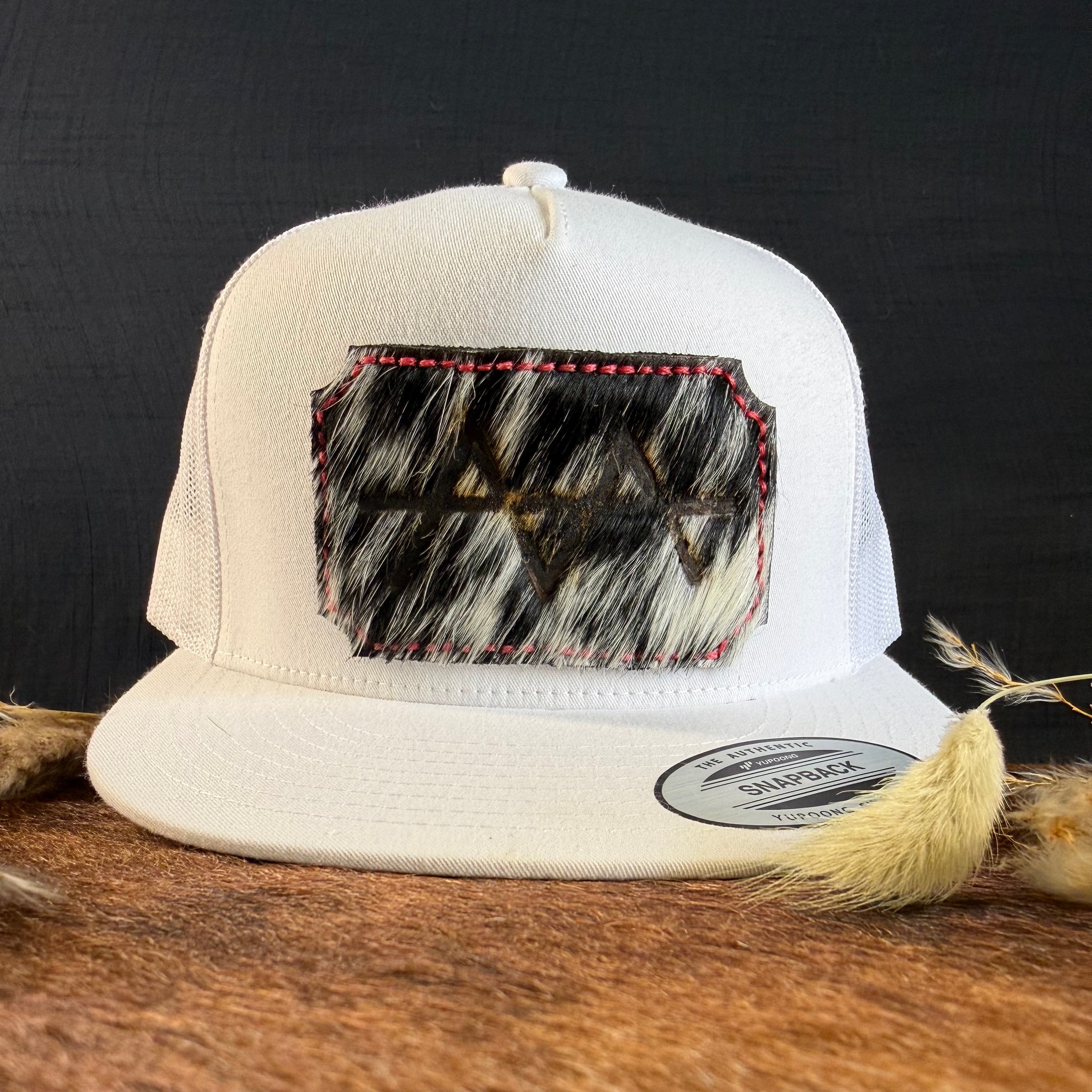 Cream snapback hat with black and white speckled cowhide patch and hot pink hand-stitched border, branded with a registered cattle brand