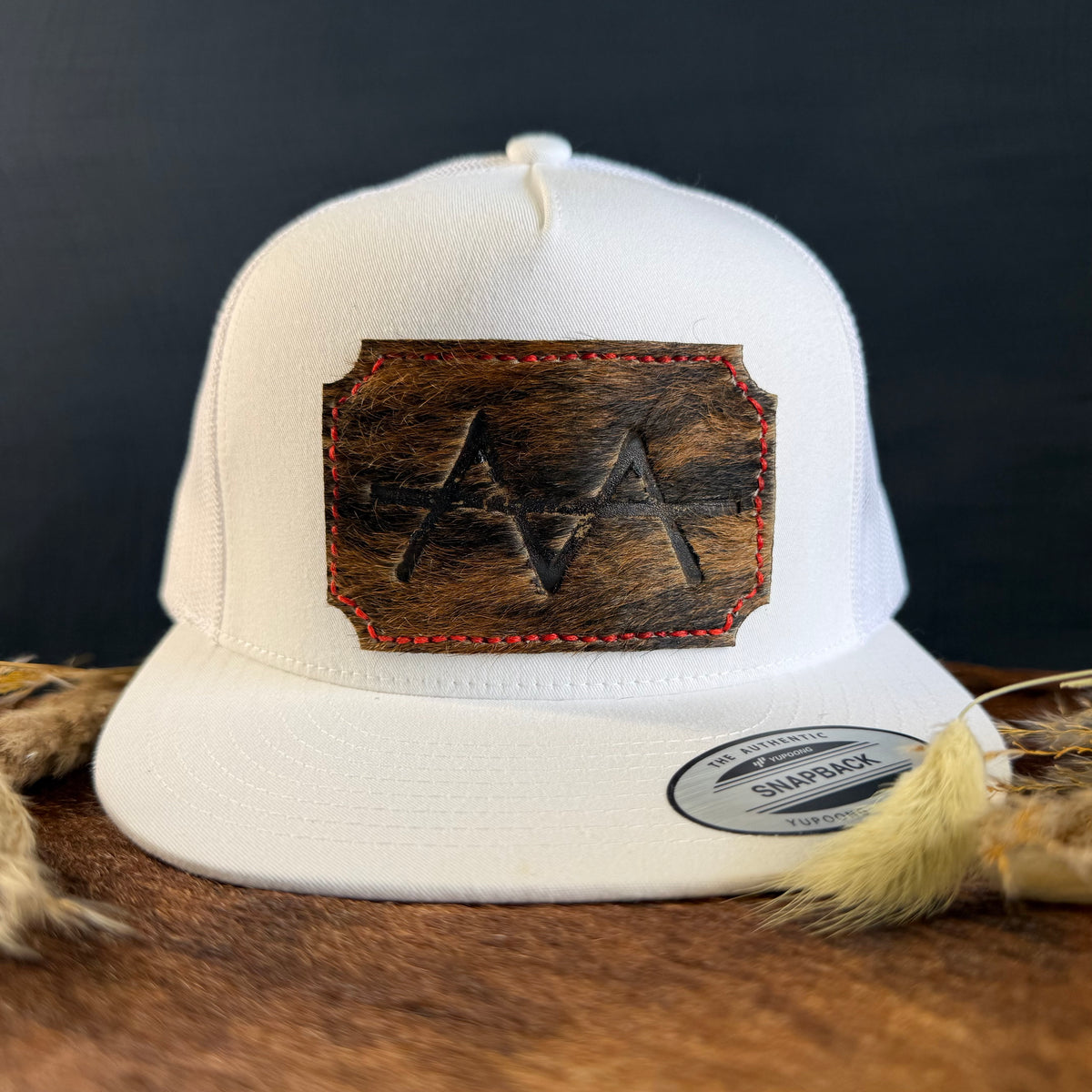 White snapback hat with scalloped espresso cowhide patch and red hand-stitched border, branded with a registered cattle brand