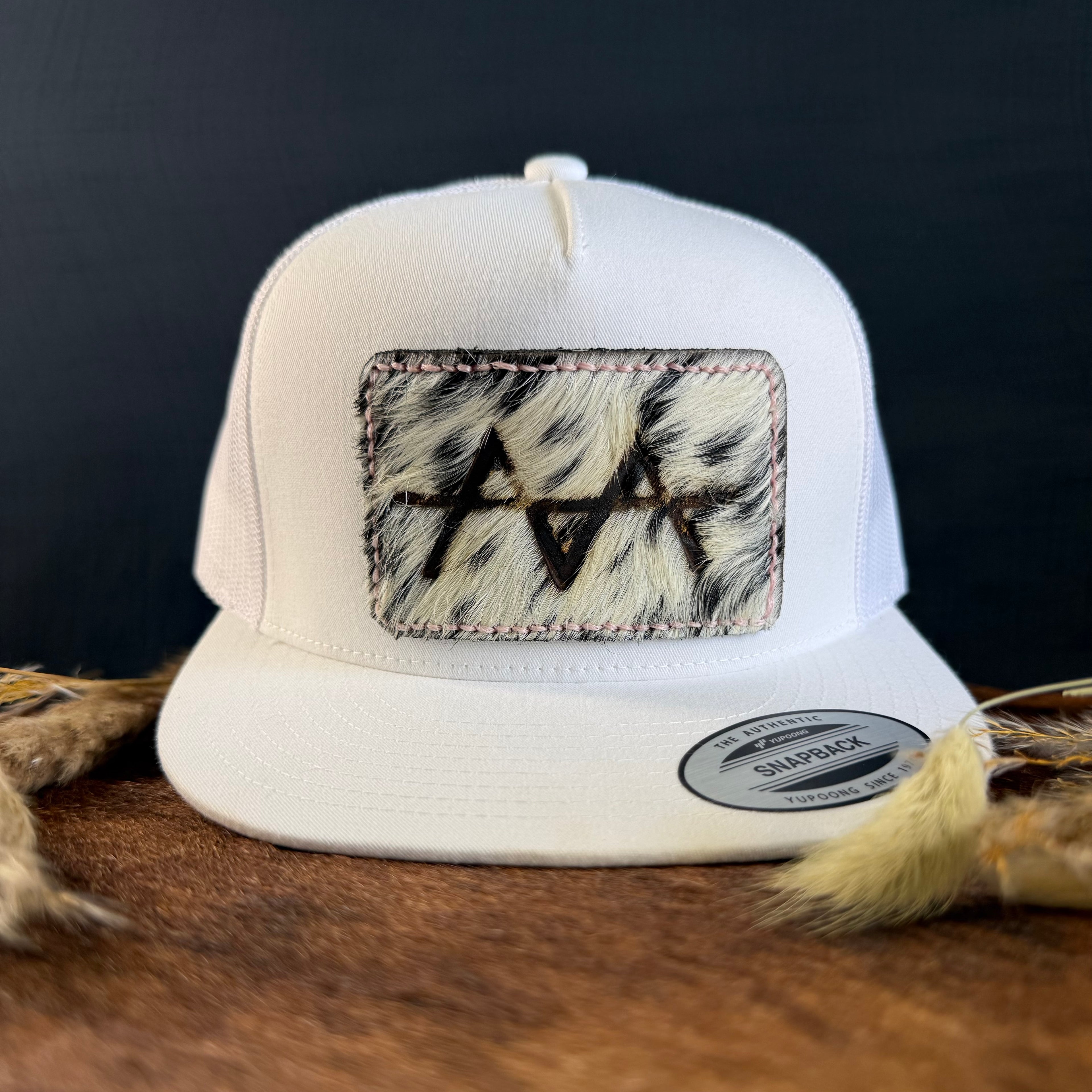 White snapback hat with black and white speckled cowhide patch and tonal stitched border, branded with a registered cattle brand