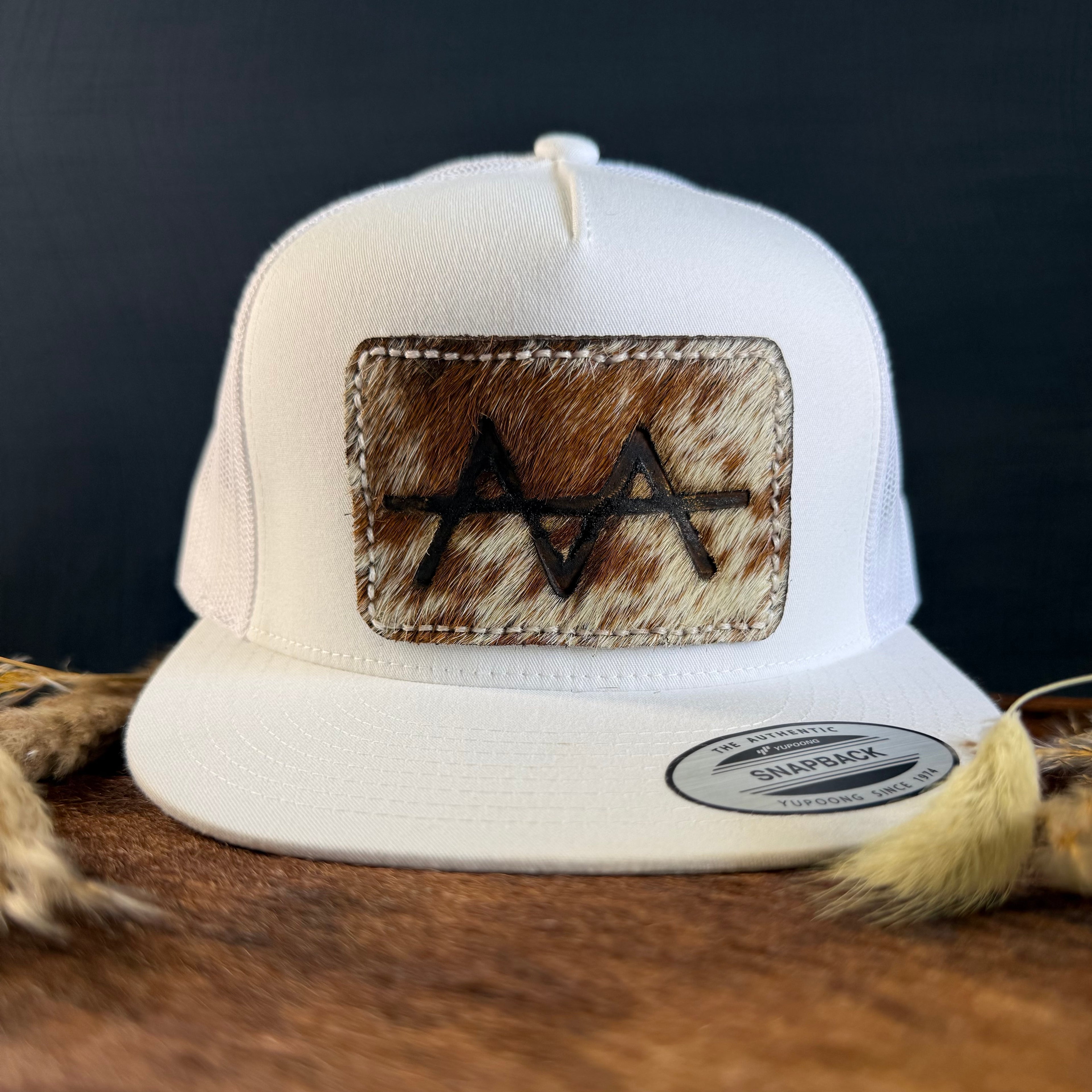 White snapback hat with mottled tan and brown cowhide patch and cream tonal stitched border, branded with a registered cattle brand