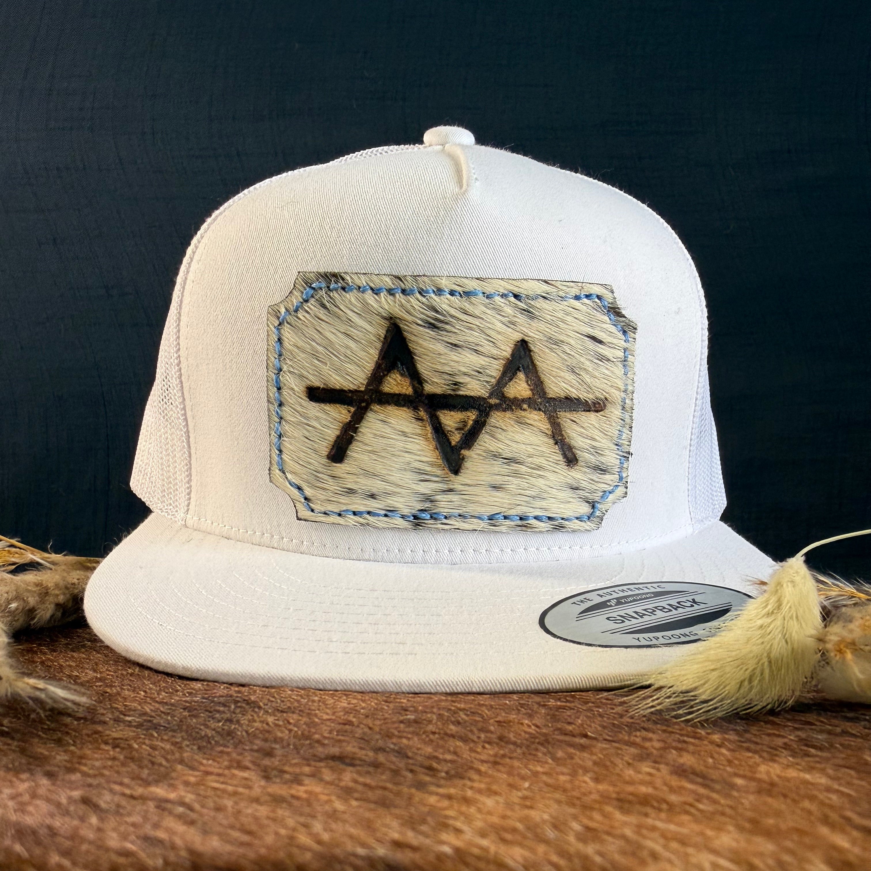 Cream snapback hat with pale ghost-speckled cowhide patch and periwinkle blue hand-stitched border, branded with a registered cattle brand