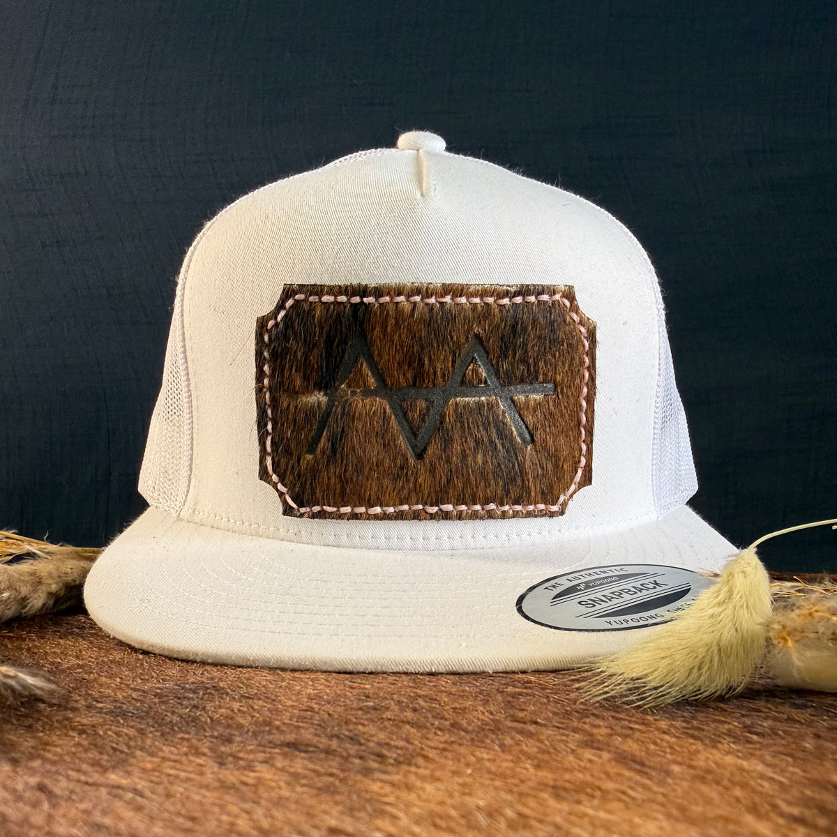 Cream snapback hat with deep espresso cowhide patch and blush pink hand-stitched border, branded with a registered cattle brand
