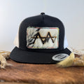 Black and white trucker snapback hat with speckled cowhide patch and multicolor rainbow hand-stitched border, branded with a registered cattle brand