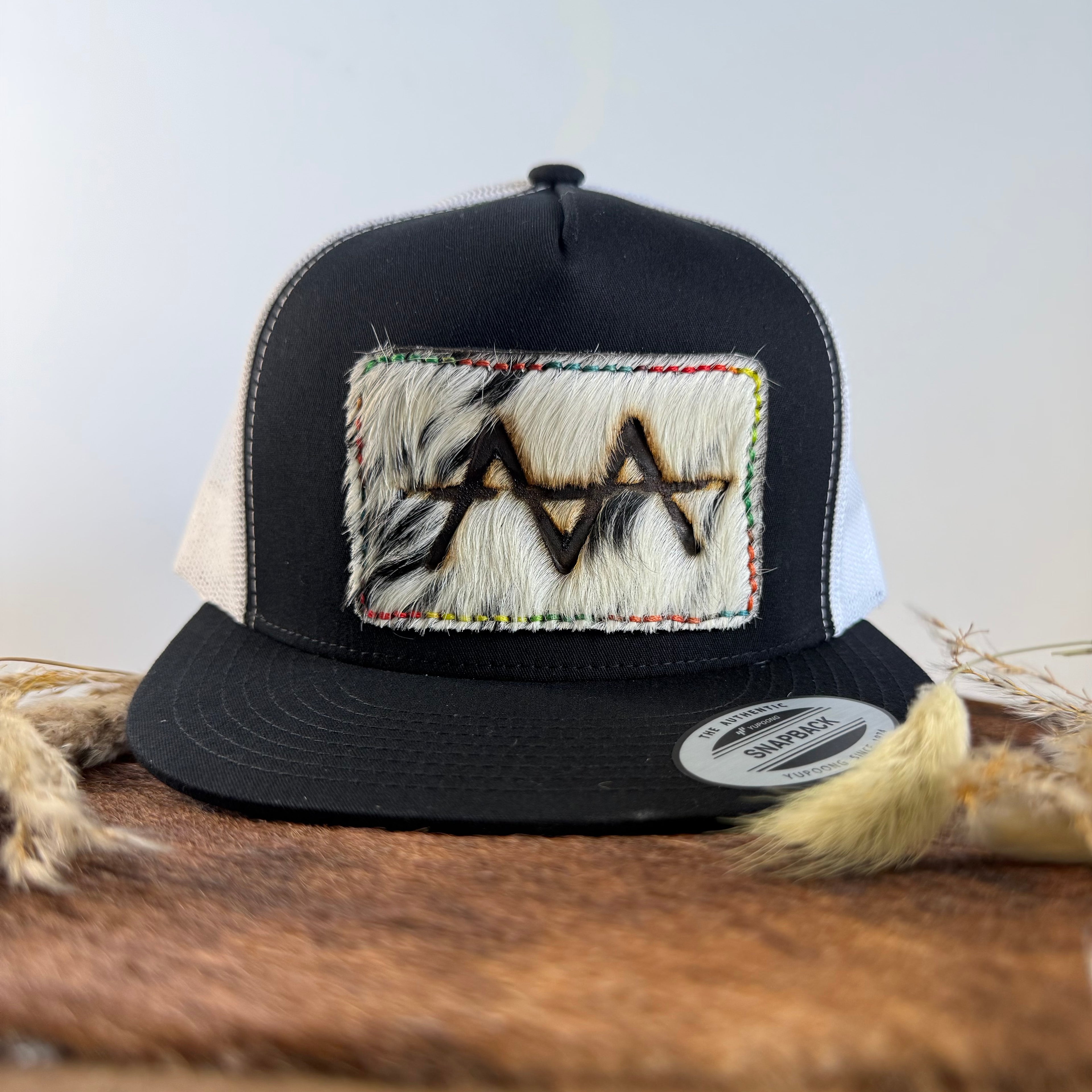 Black and white trucker snapback hat with speckled cowhide patch and multicolor rainbow hand-stitched border, branded with a registered cattle brand