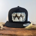 Black and white trucker snapback hat with ghost-speckled cowhide patch and blue hand-stitched border, branded with a registered cattle brand