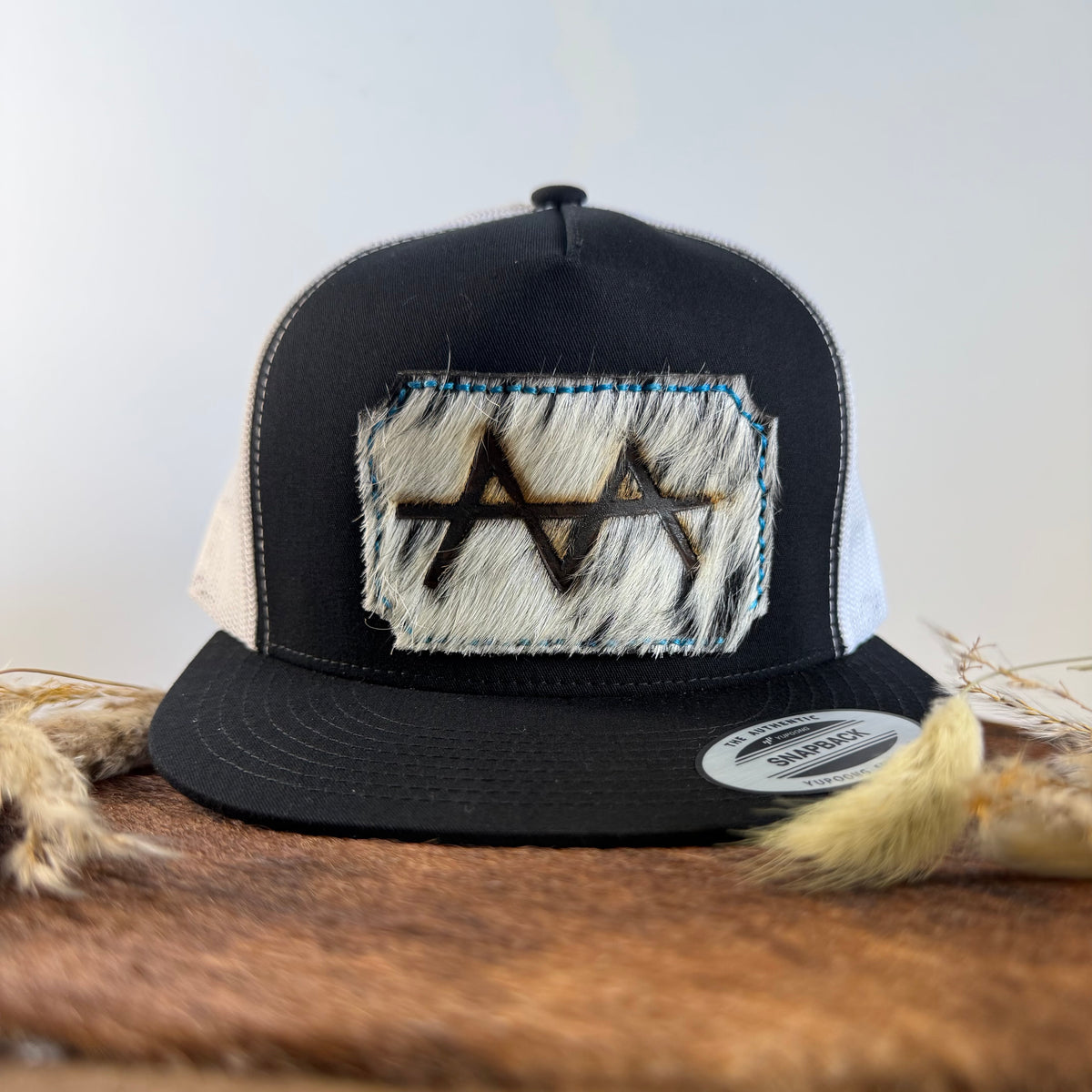 Black and white trucker snapback hat with ghost-speckled cowhide patch and blue hand-stitched border, branded with a registered cattle brand