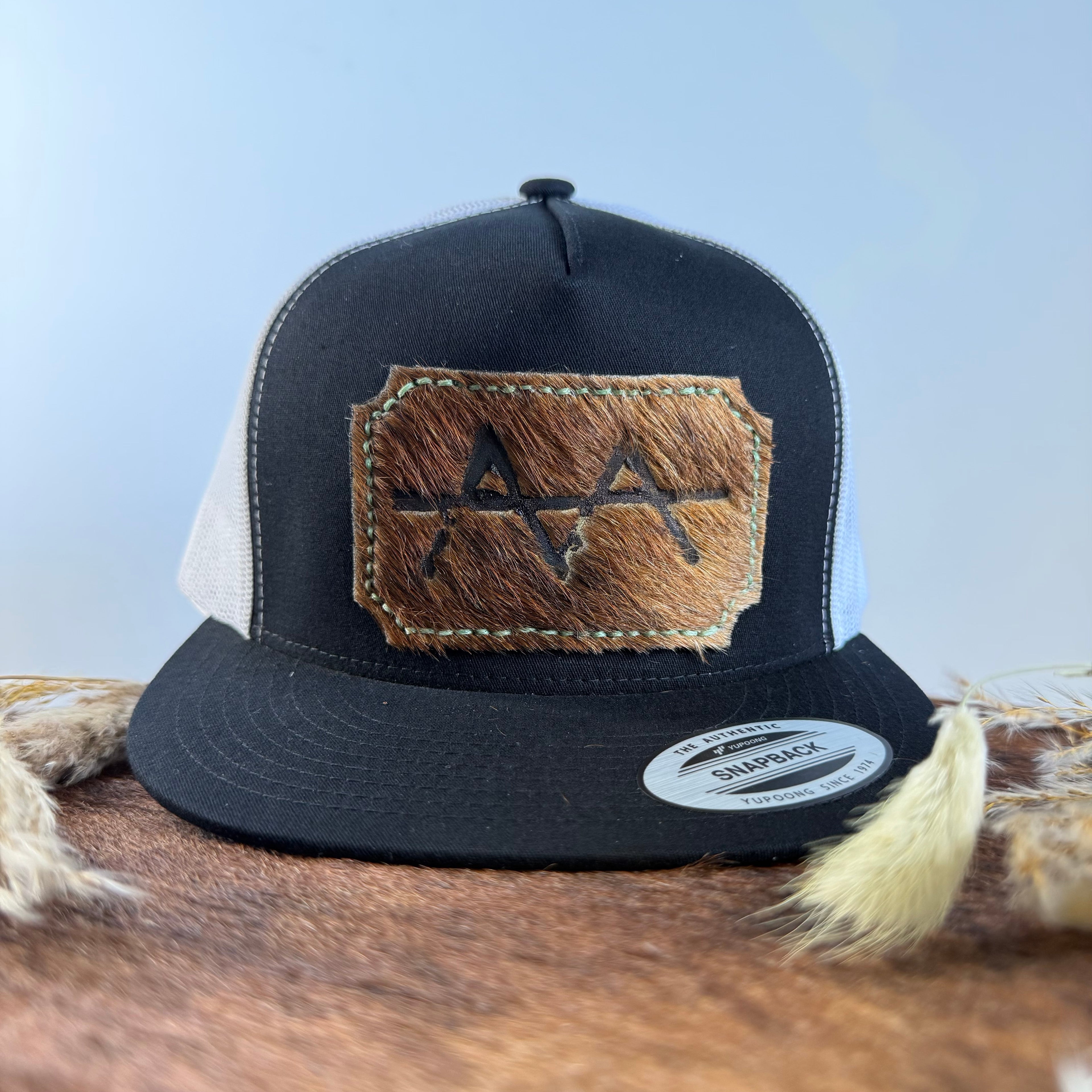Black and white trucker snapback hat with scalloped espresso cowhide patch and olive gold tonal stitched border, branded with a registered cattle brand