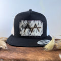 Black and white trucker snapback hat with pale ghost-speckled cowhide patch and tonal white stitched border, branded with a registered cattle brand