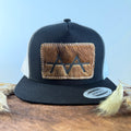 Black and white trucker snapback hat with warm brown cowhide patch and gold tonal stitched border, branded with a registered cattle brand