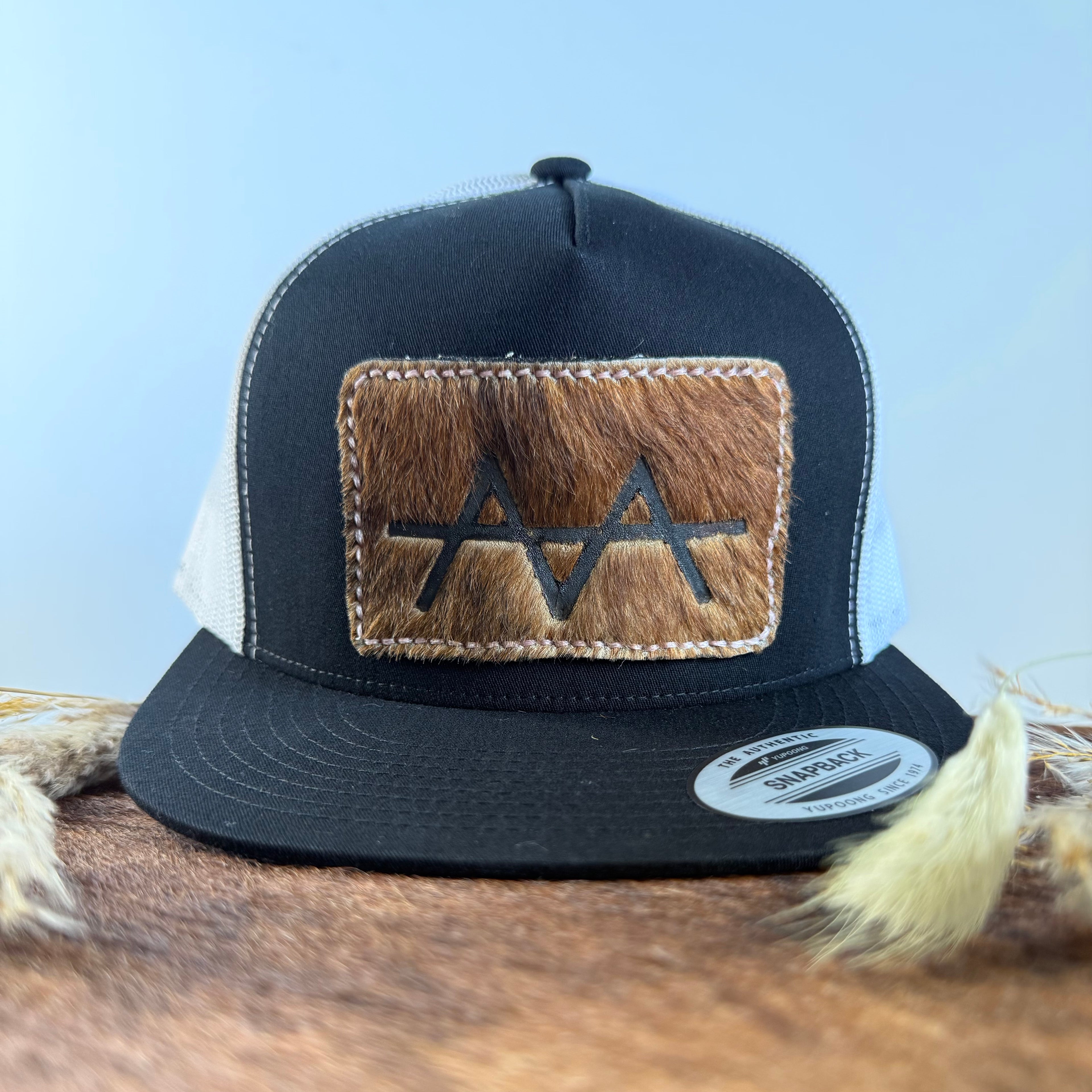 Black and white trucker snapback hat with warm brown cowhide patch and gold tonal stitched border, branded with a registered cattle brand