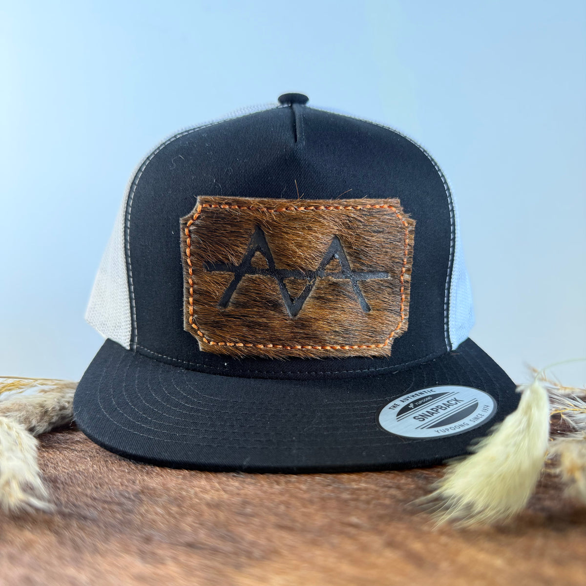 Black and white trucker snapback hat with scalloped warm brown cowhide patch and orange hand-stitched border, branded with a registered cattle brand