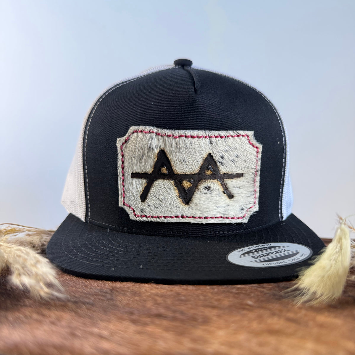 Black and white trucker snapback hat with pale ghost-speckled cowhide patch and red hand-stitched border, branded with a registered cattle brand