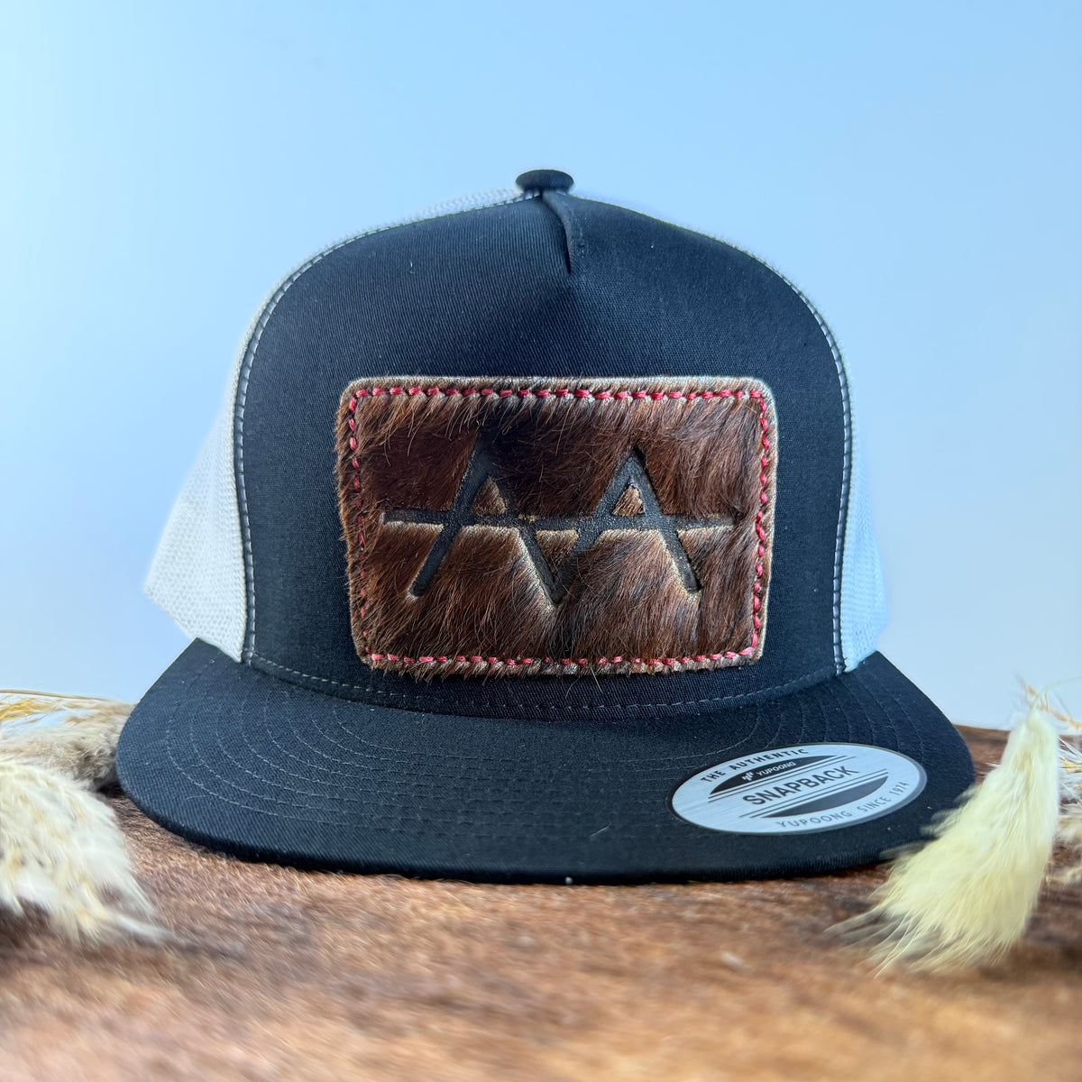 Black and white trucker snapback hat with deep espresso cowhide patch and red hand-stitched border, branded with a registered cattle brand