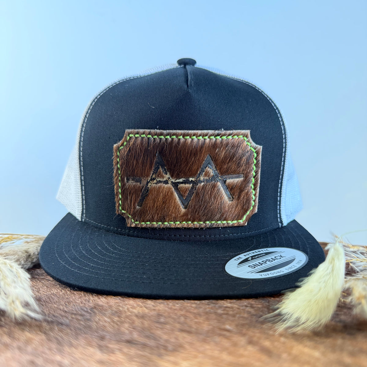 Black and white trucker snapback hat with scalloped warm brown cowhide patch and neon yellow-green hand-stitched border, branded with a registered cattle brand