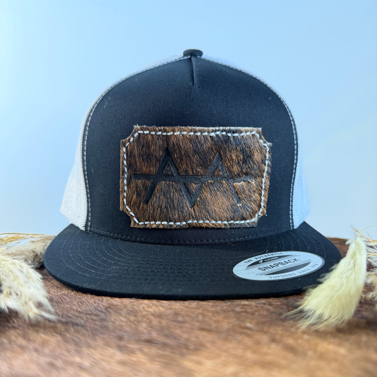 Black and white trucker snapback hat with scalloped dark brindle cowhide patch and cream tonal stitched border, branded with a registered cattle brand
