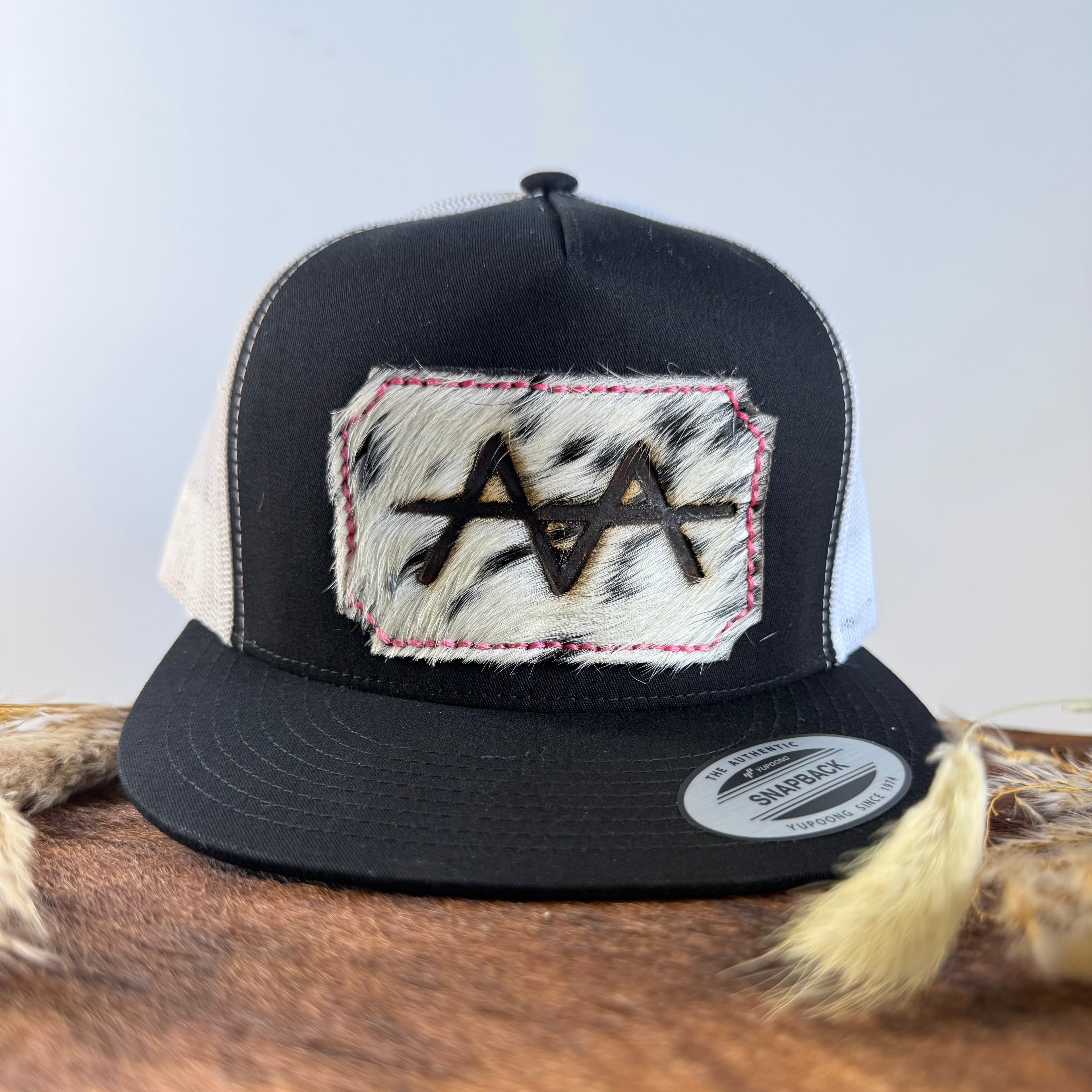 Black and white trucker snapback hat with bold black and white speckled cowhide patch and pink hand-stitched border, branded with a registered cattle brand
