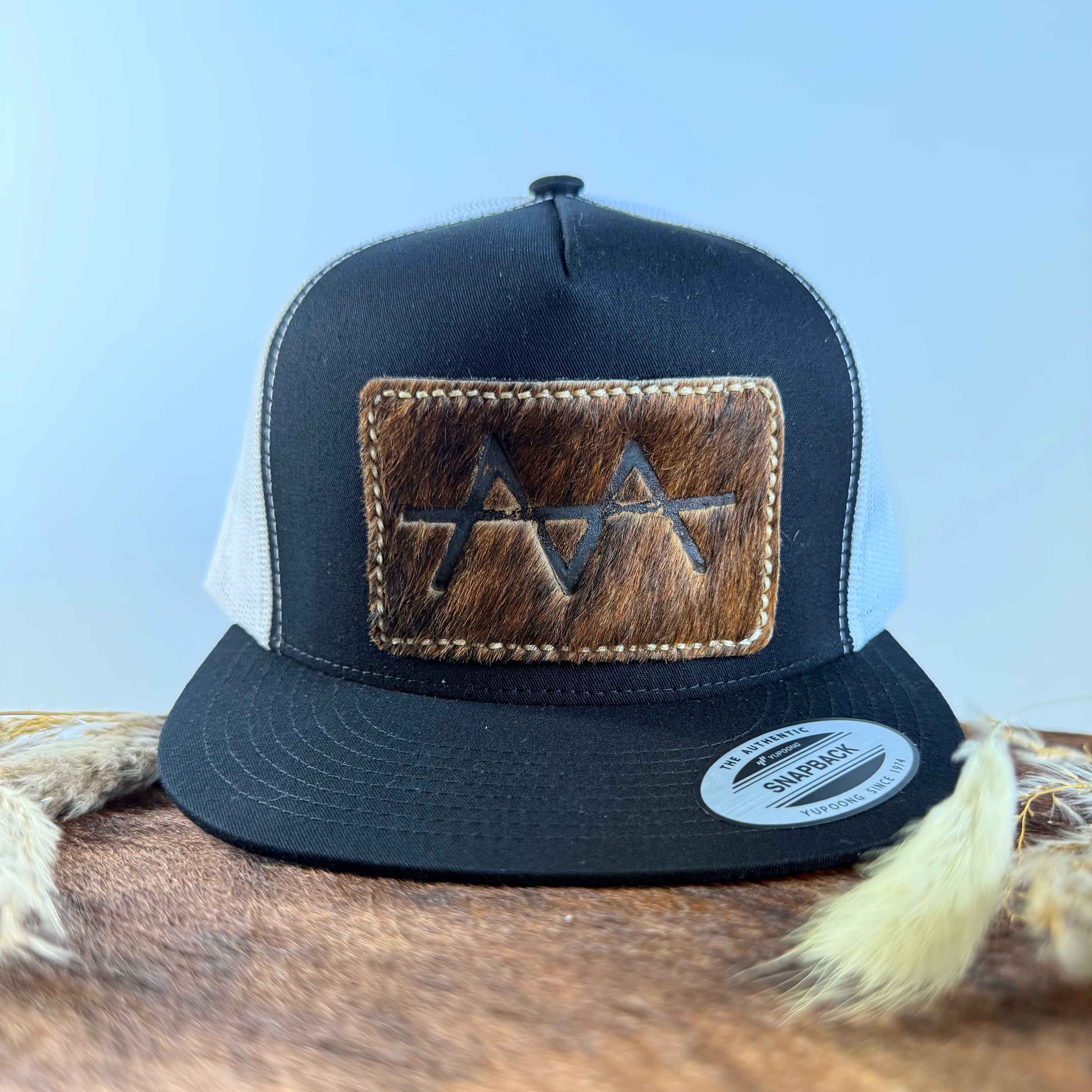Black and white trucker snapback hat with dark brindle cowhide patch and tan tonal stitched border, branded with a registered cattle brand