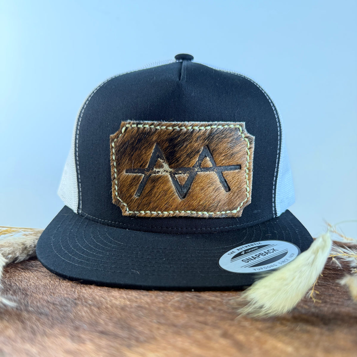 Black and white trucker snapback hat with scalloped medium brown brindle cowhide patch and cream tonal stitched border, branded with a registered cattle brand