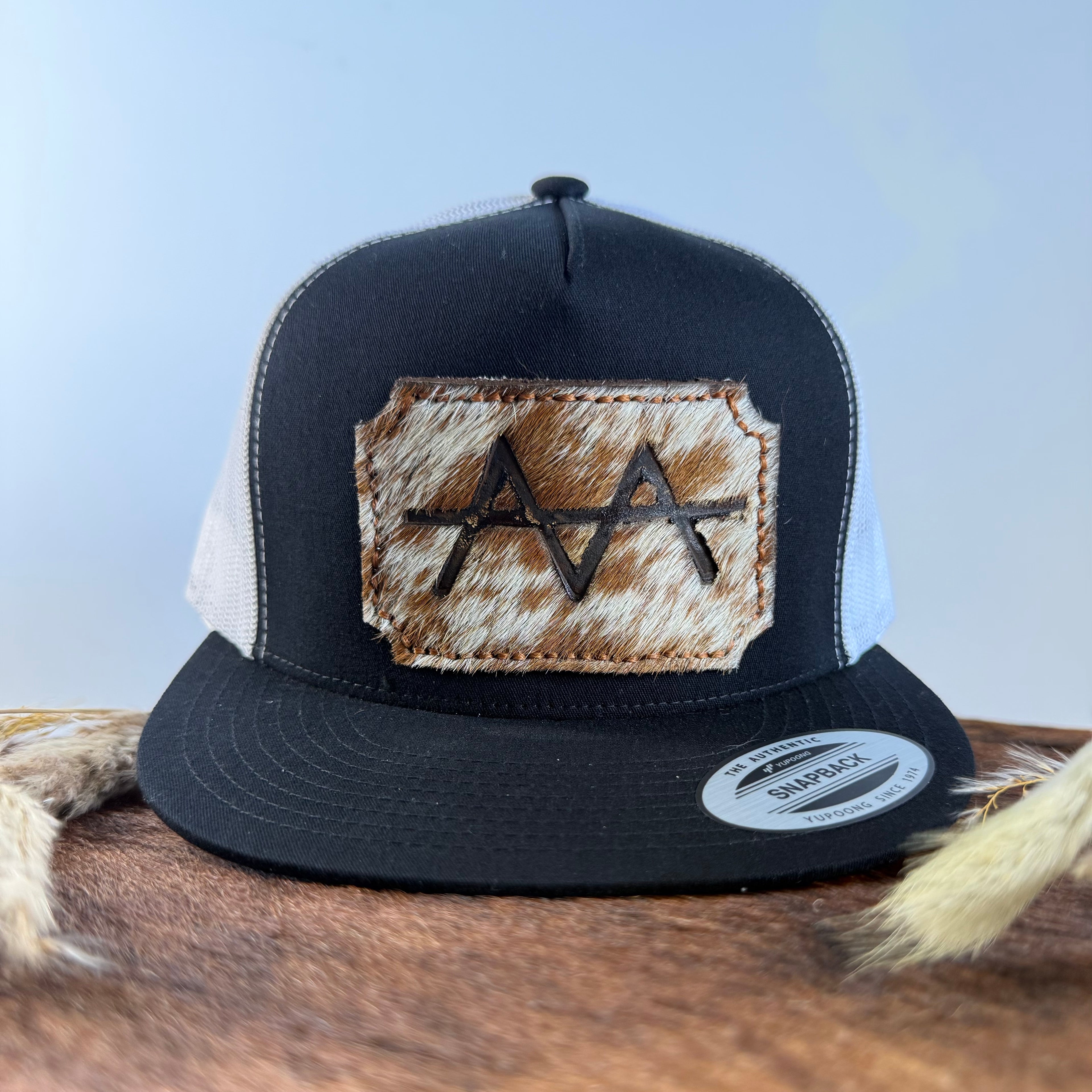Black and white trucker snapback hat with scalloped champagne speckled cowhide patch and tonal stitched border, branded with a registered cattle brand