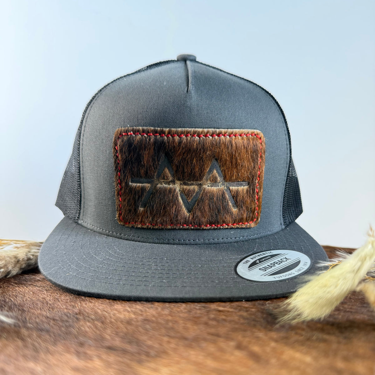 Charcoal grey snapback hat with dark brindle cowhide patch and red hand-stitched border, branded with a registered cattle brand
