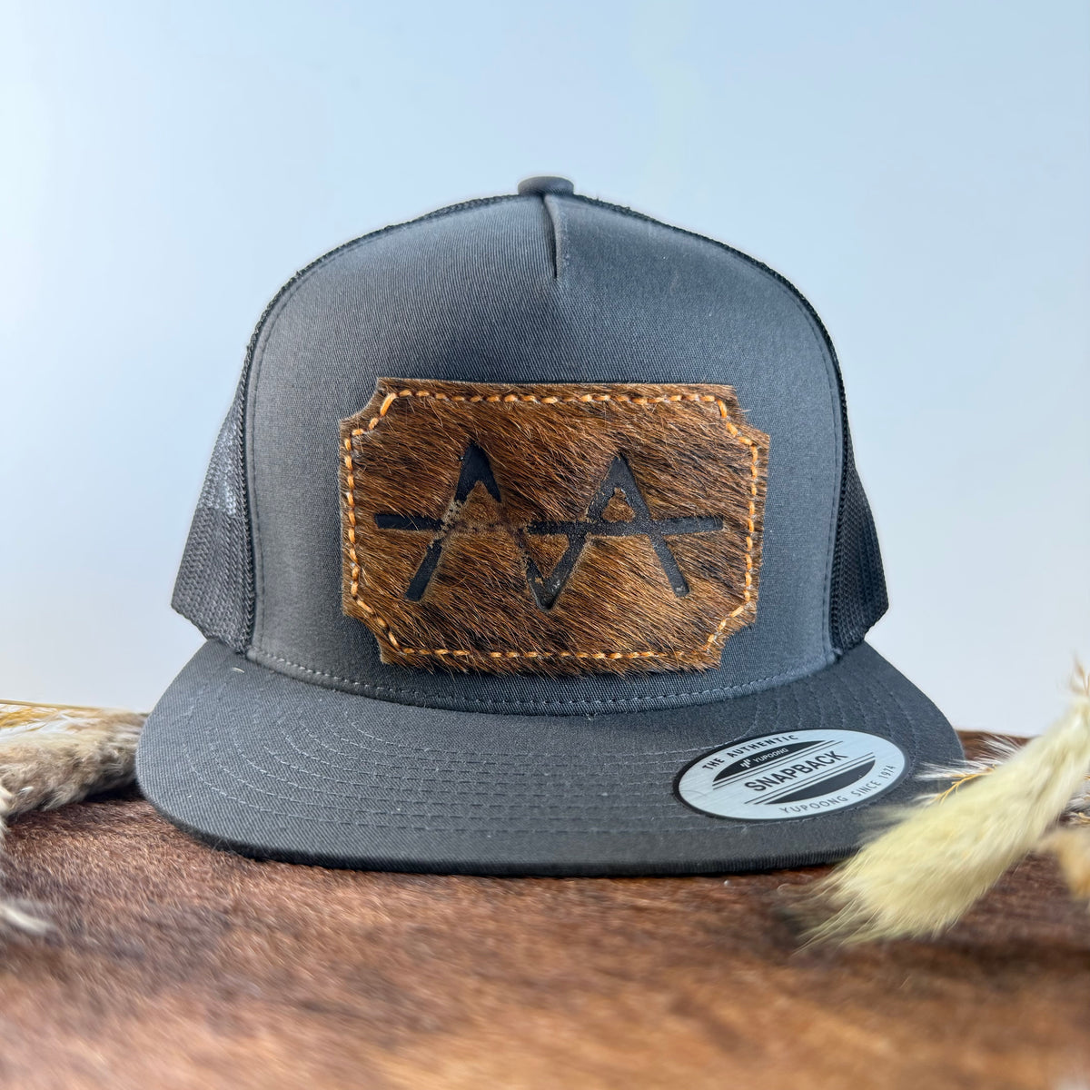 Charcoal grey snapback hat with scalloped golden brindle cowhide patch and gold tonal stitched border, branded with a registered cattle brand