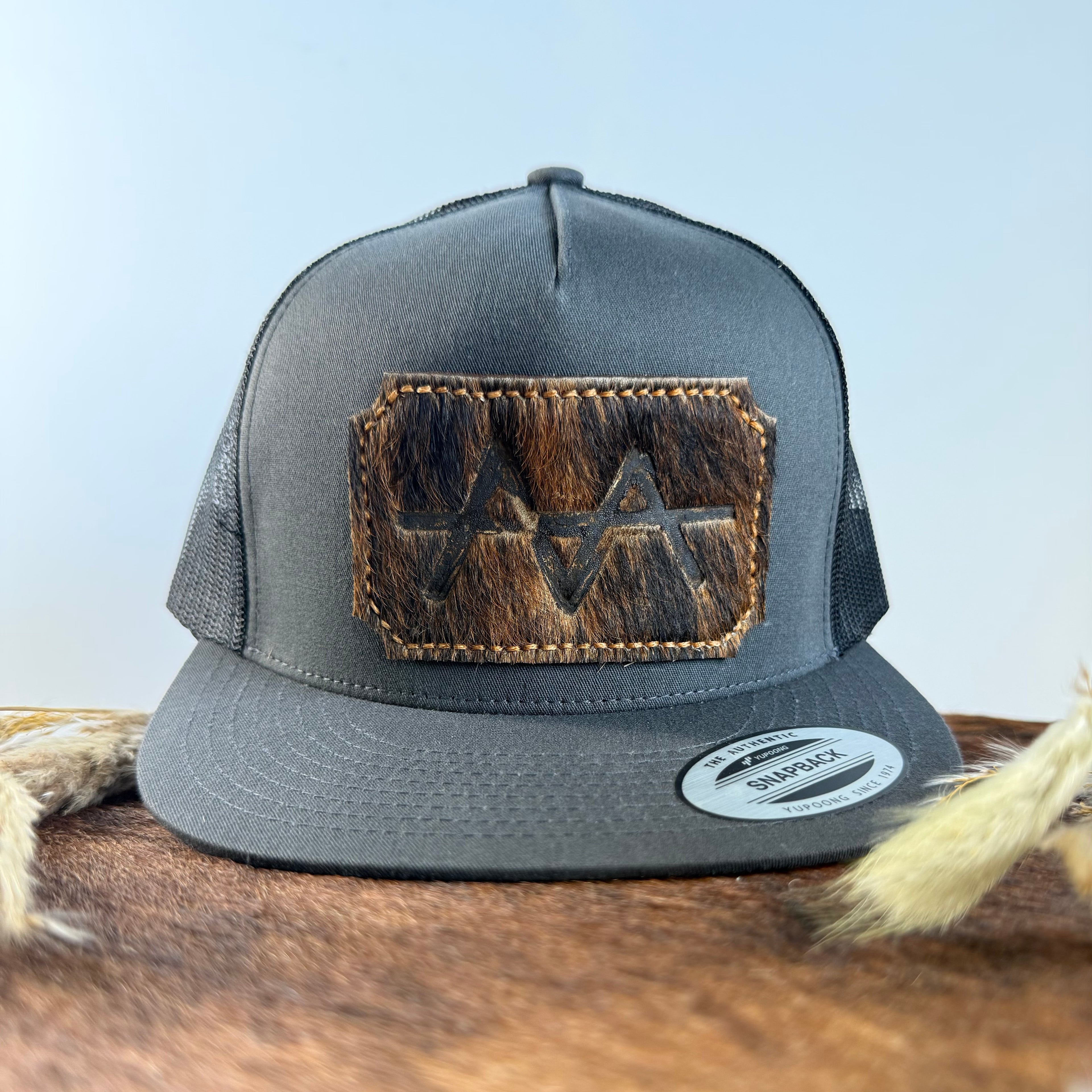 Charcoal grey snapback hat with near-black brindle cowhide patch and gold tonal stitched border, branded with a registered cattle brand
