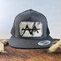 Charcoal grey snapback hat with bold black and white speckled cowhide patch and tonal stitched border, branded with a registered cattle brand