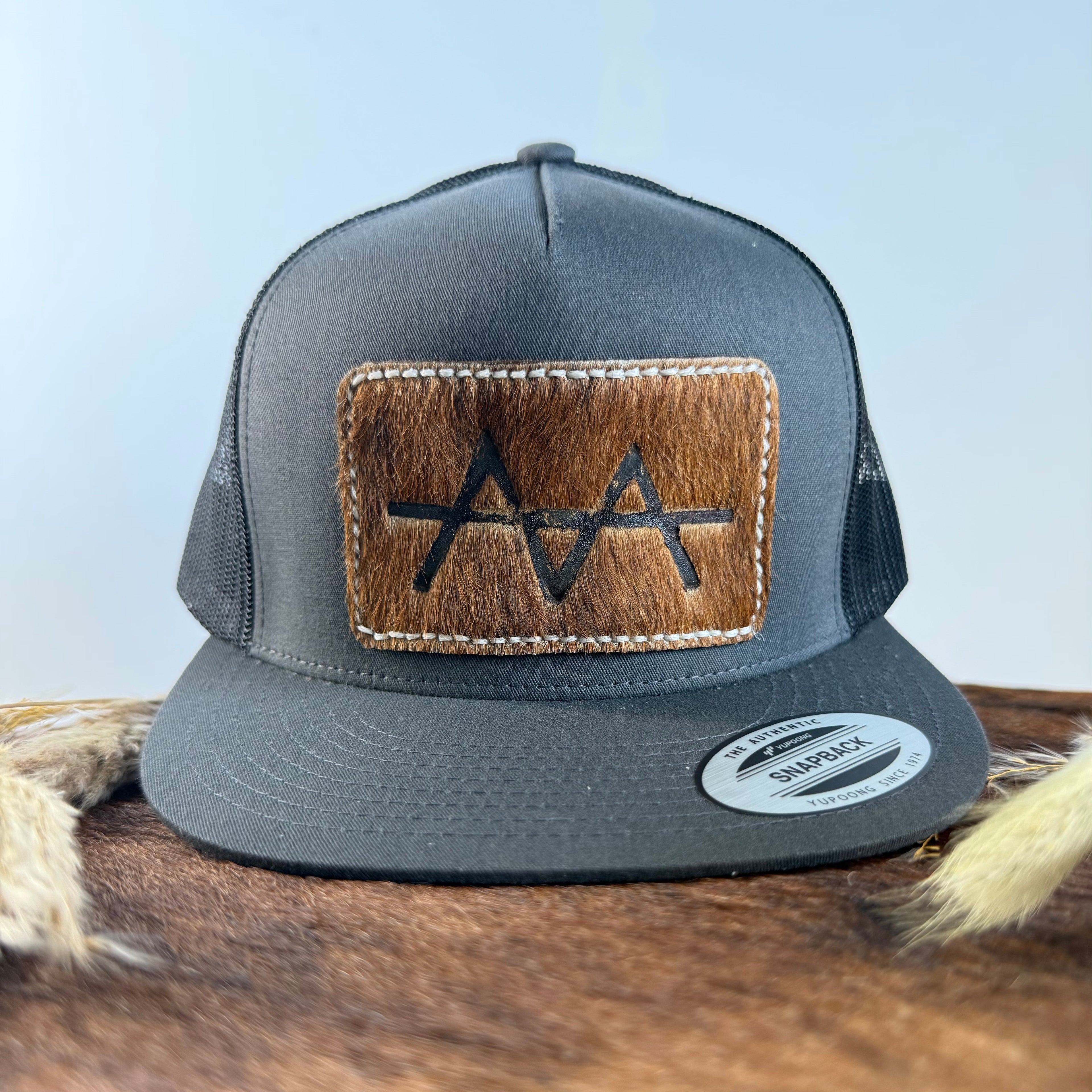 Charcoal grey snapback hat with warm brown cowhide patch and cream tonal stitched border, branded with a registered cattle brand