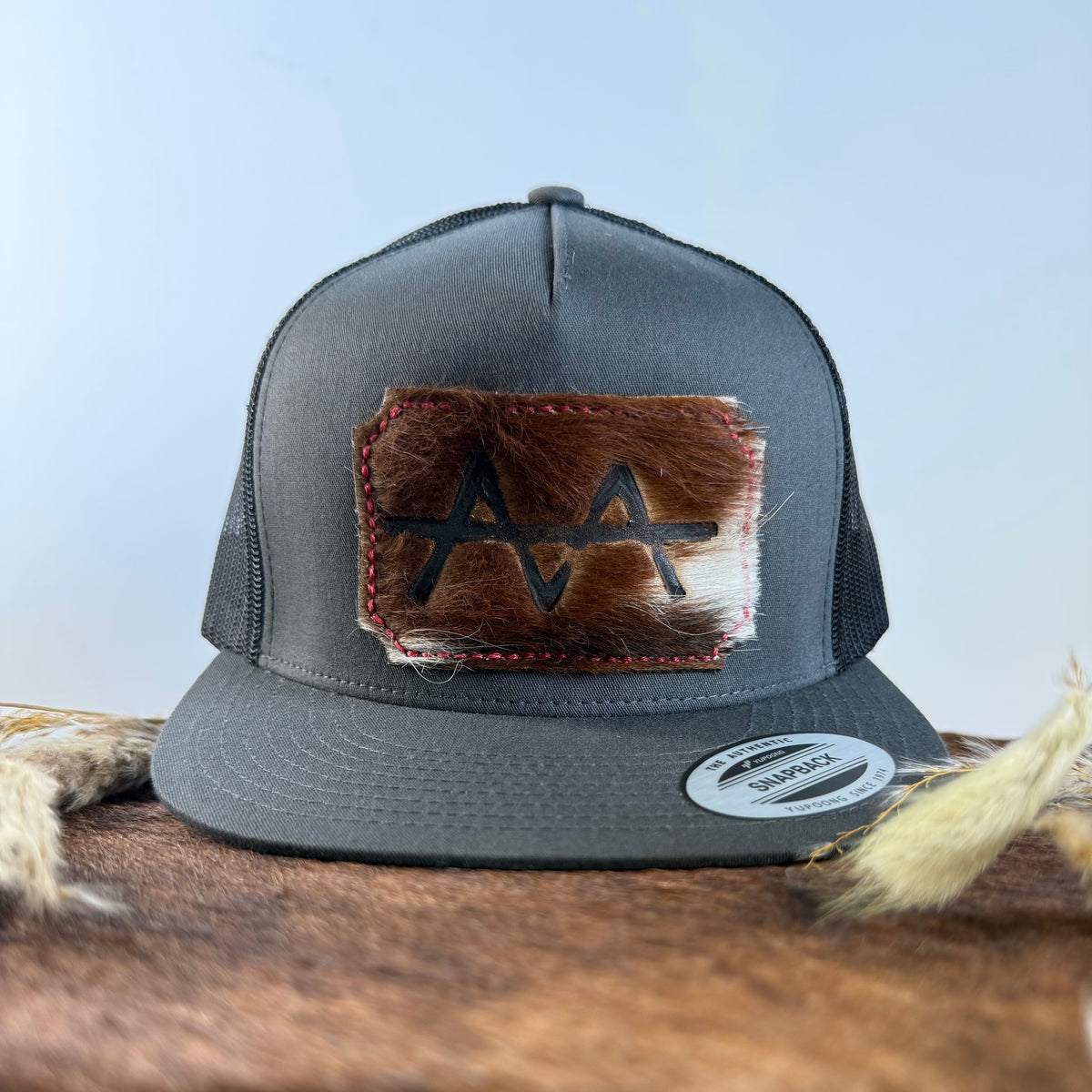 Charcoal grey snapback hat with two-tone brown and cream cowhide patch and red hand-stitched border, branded with a registered cattle brand