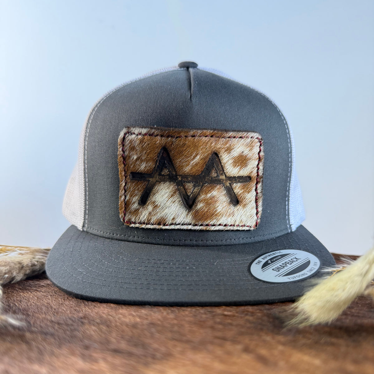 Charcoal and white trucker snapback hat with golden speckled cowhide patch and tonal stitched border, branded with a registered cattle brand