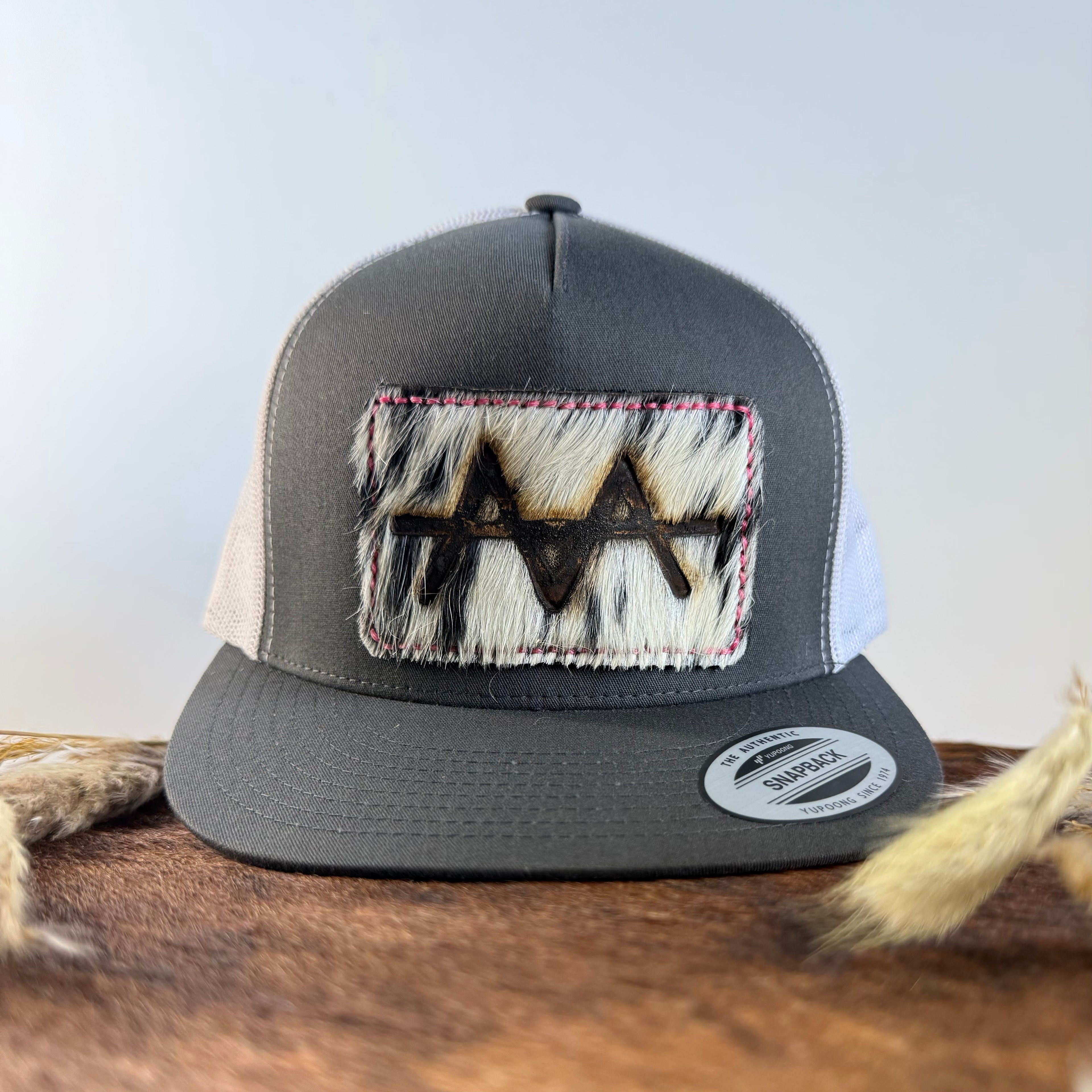 Charcoal and white trucker snapback hat with bold black and white speckled cowhide patch and pink hand-stitched border, branded with a registered cattle brand