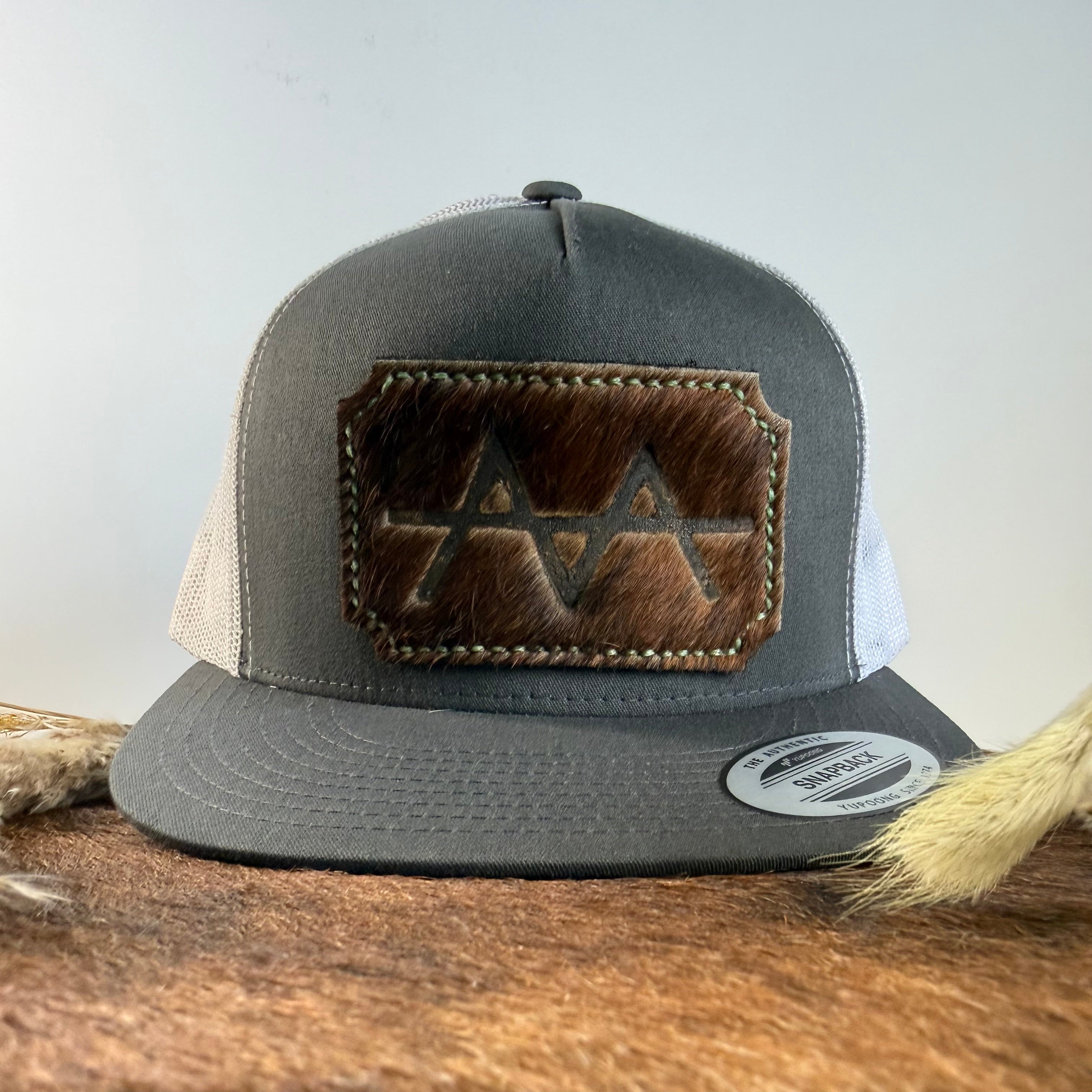 Charcoal and white trucker snapback hat with scalloped espresso brindle cowhide patch and tonal stitched border, branded with a registered cattle brand