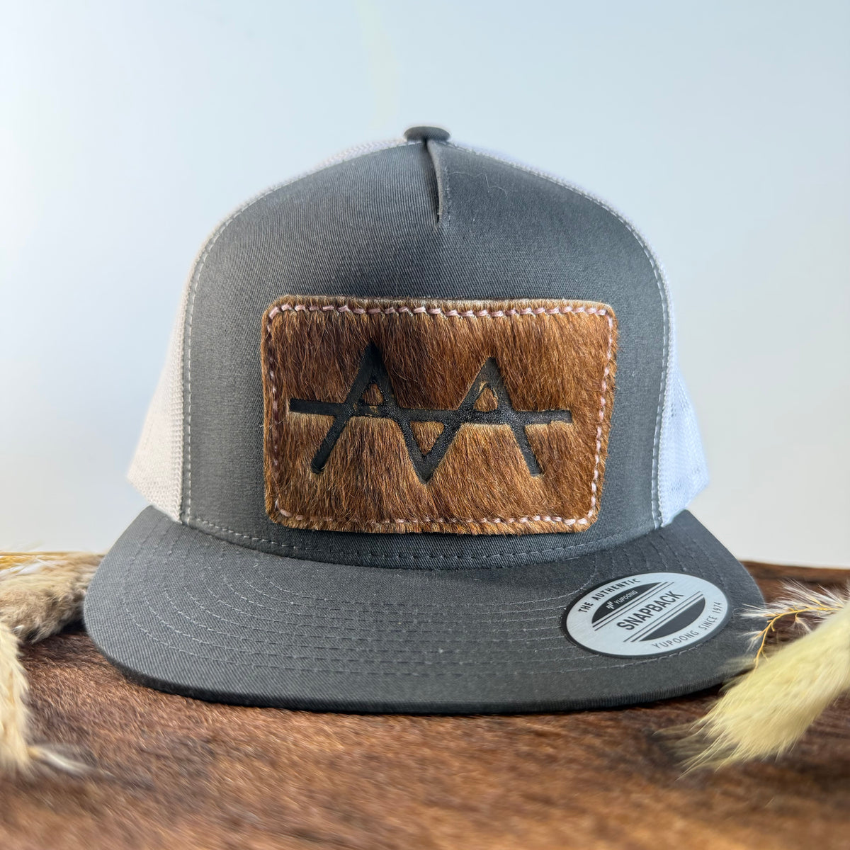 Charcoal and white trucker snapback hat with golden brown cowhide patch and blush rose hand-stitched border, branded with a registered cattle brand