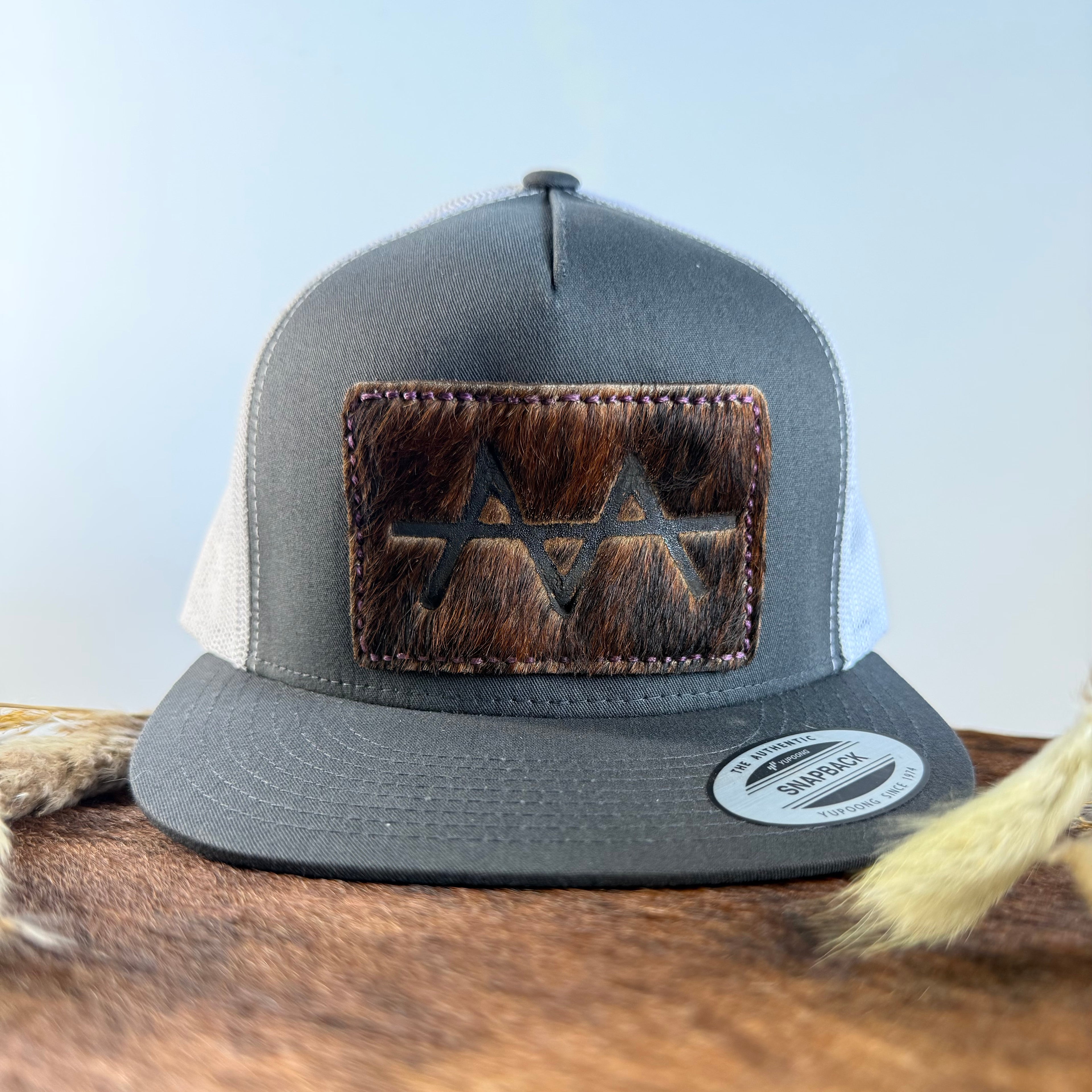 Charcoal and white trucker snapback hat with deep dark brindle cowhide patch and burgundy wine hand-stitched border, branded with a registered cattle brand