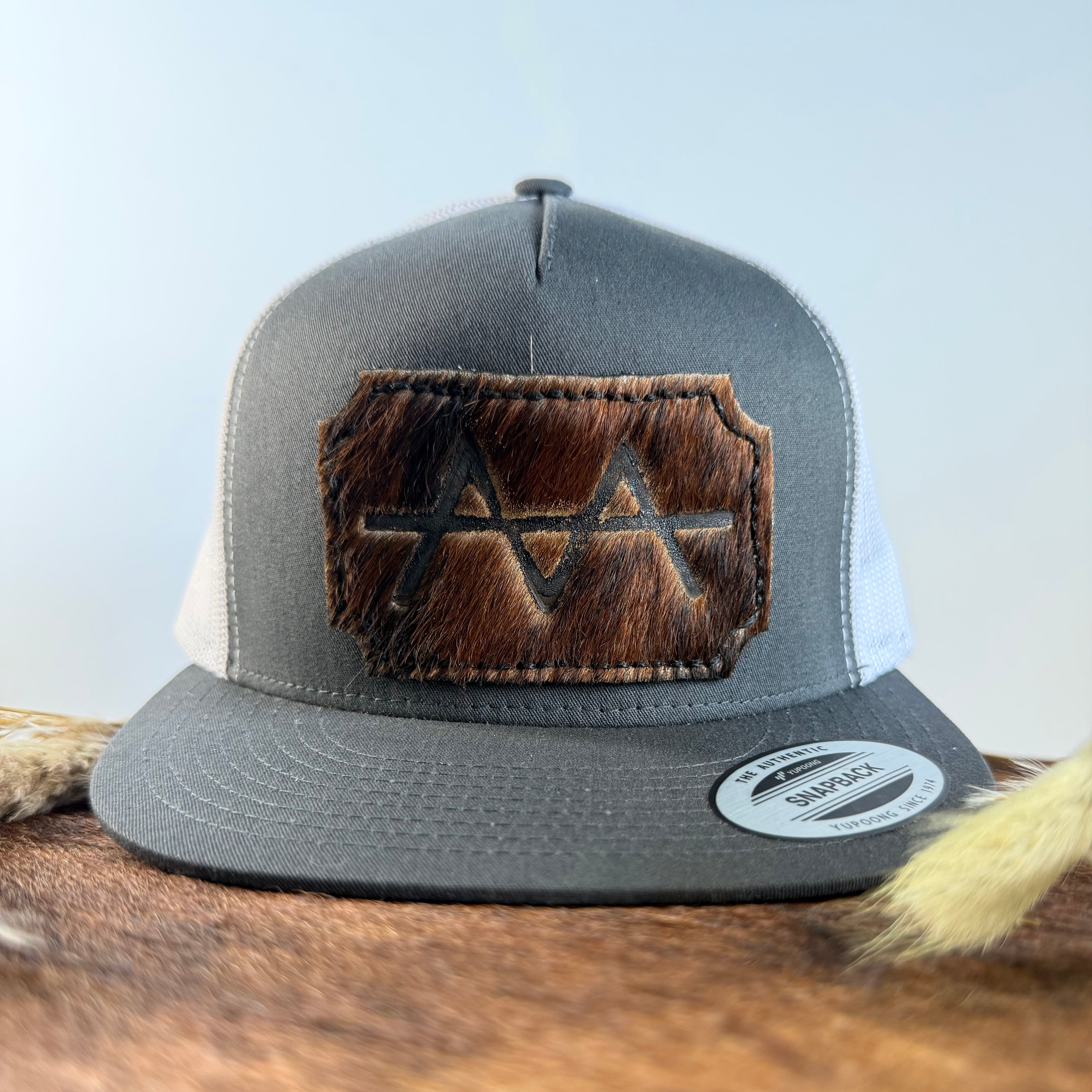 Charcoal and white trucker snapback hat with scalloped dark brindle cowhide patch and tonal dark stitched border, branded with a registered cattle brand