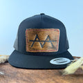 All-black trucker snapback hat with golden brown cowhide patch and red hand-stitched border, branded with a registered cattle brand