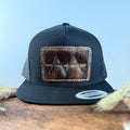 All-black trucker snapback hat with deep dark brindle cowhide patch and cream tonal stitched border, branded with a registered cattle brand
