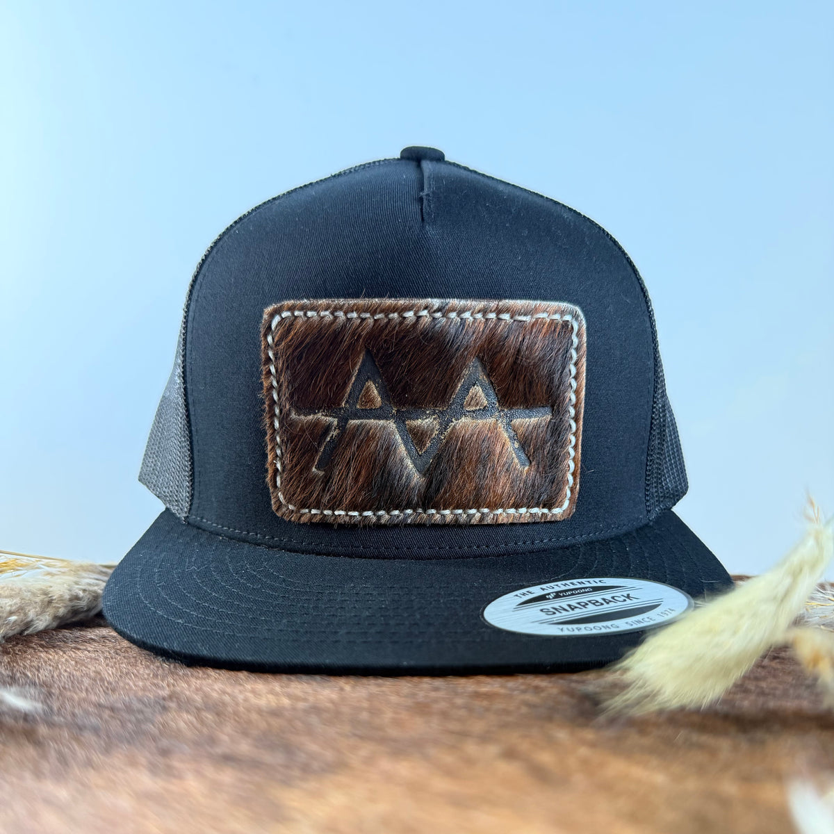 All-black trucker snapback hat with deep dark brindle cowhide patch and cream tonal stitched border, branded with a registered cattle brand