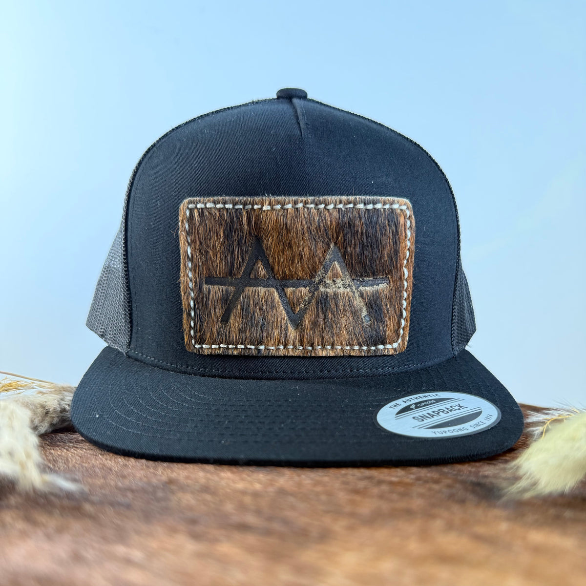 All-black trucker snapback hat with deep dark brindle cowhide patch and tan tonal stitched border, branded with a registered cattle brand