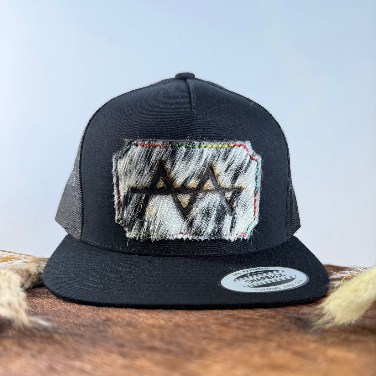 All-black trucker snapback hat with bold black and white speckled cowhide patch and multicolor rainbow hand-stitched border, branded with a registered cattle brand
