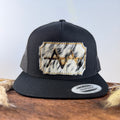 All-black trucker snapback hat with bold black and white speckled cowhide patch and gold tonal stitched border, branded with a registered cattle brand