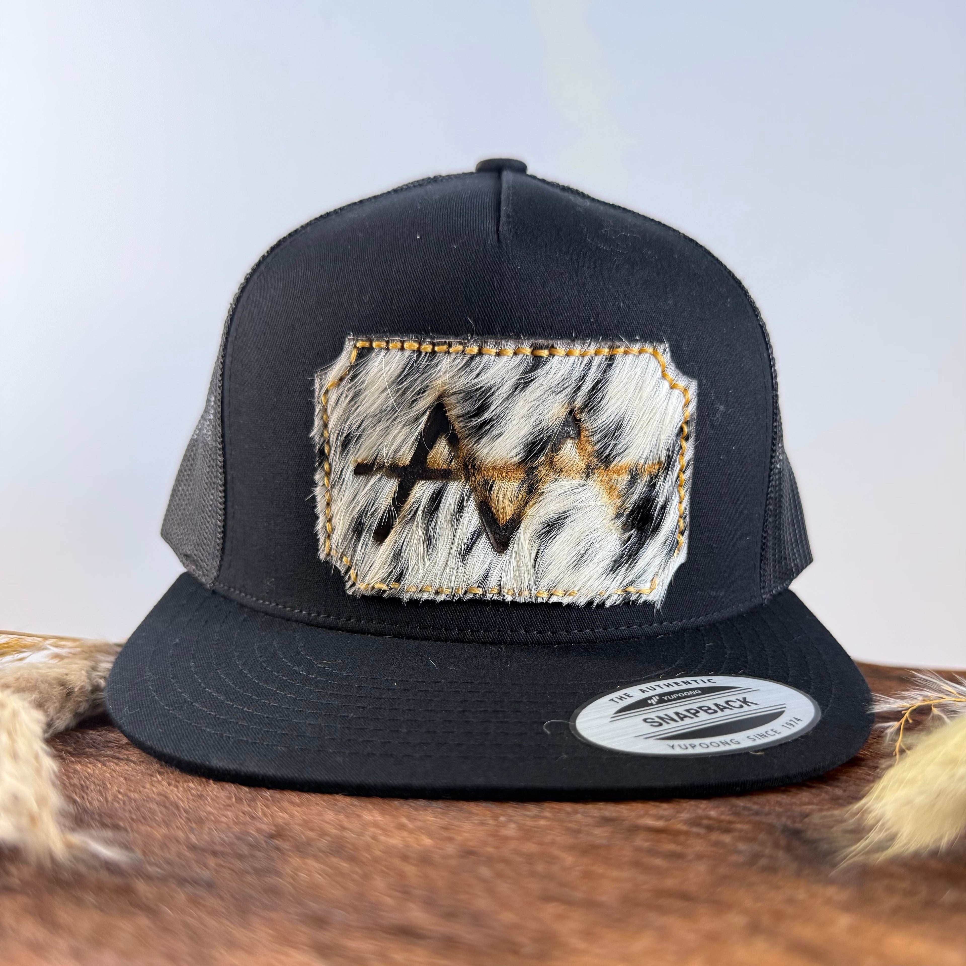All-black trucker snapback hat with bold black and white speckled cowhide patch and gold tonal stitched border, branded with a registered cattle brand