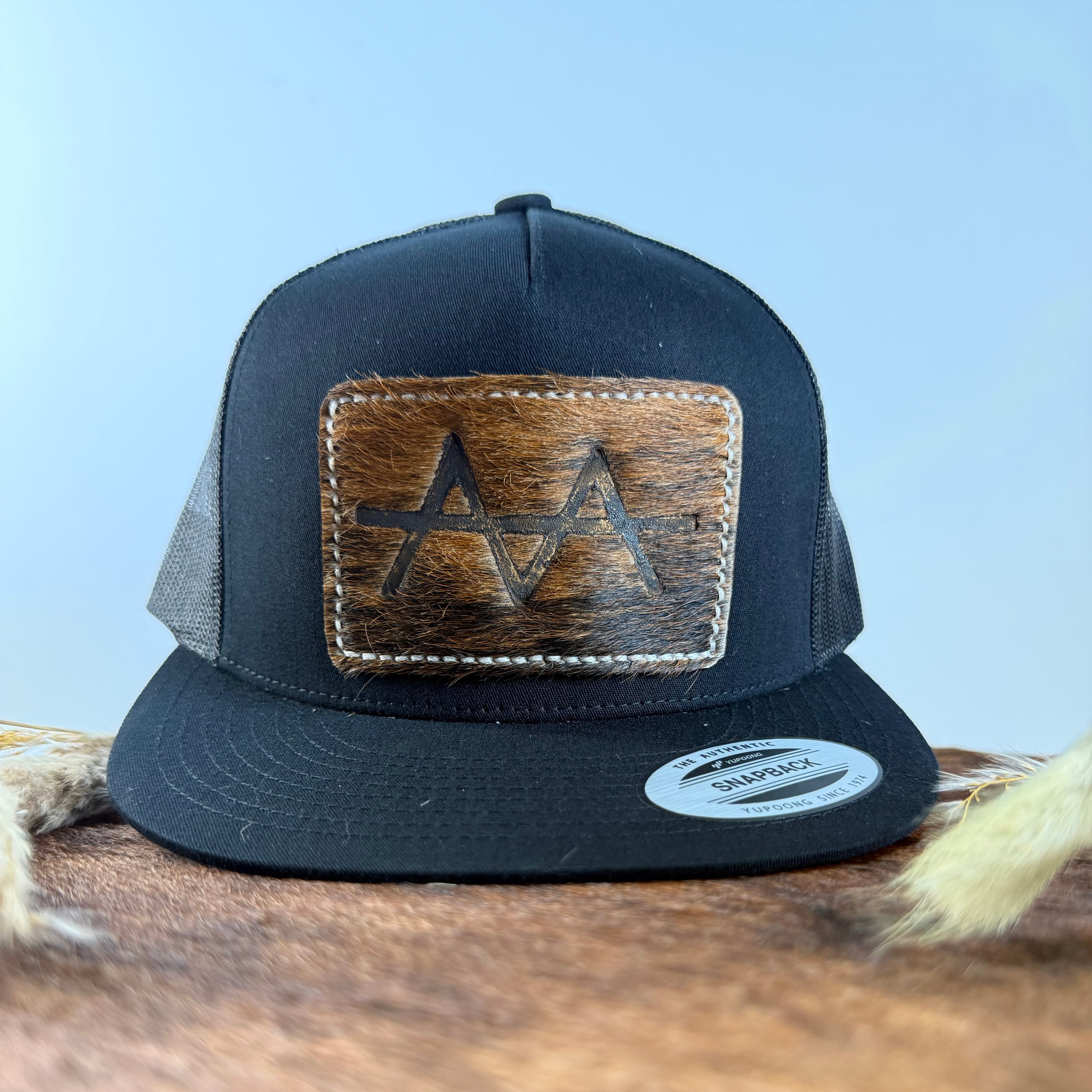 All-black trucker snapback hat with warm brindle cowhide patch and white tonal stitched border, branded with a registered cattle brand