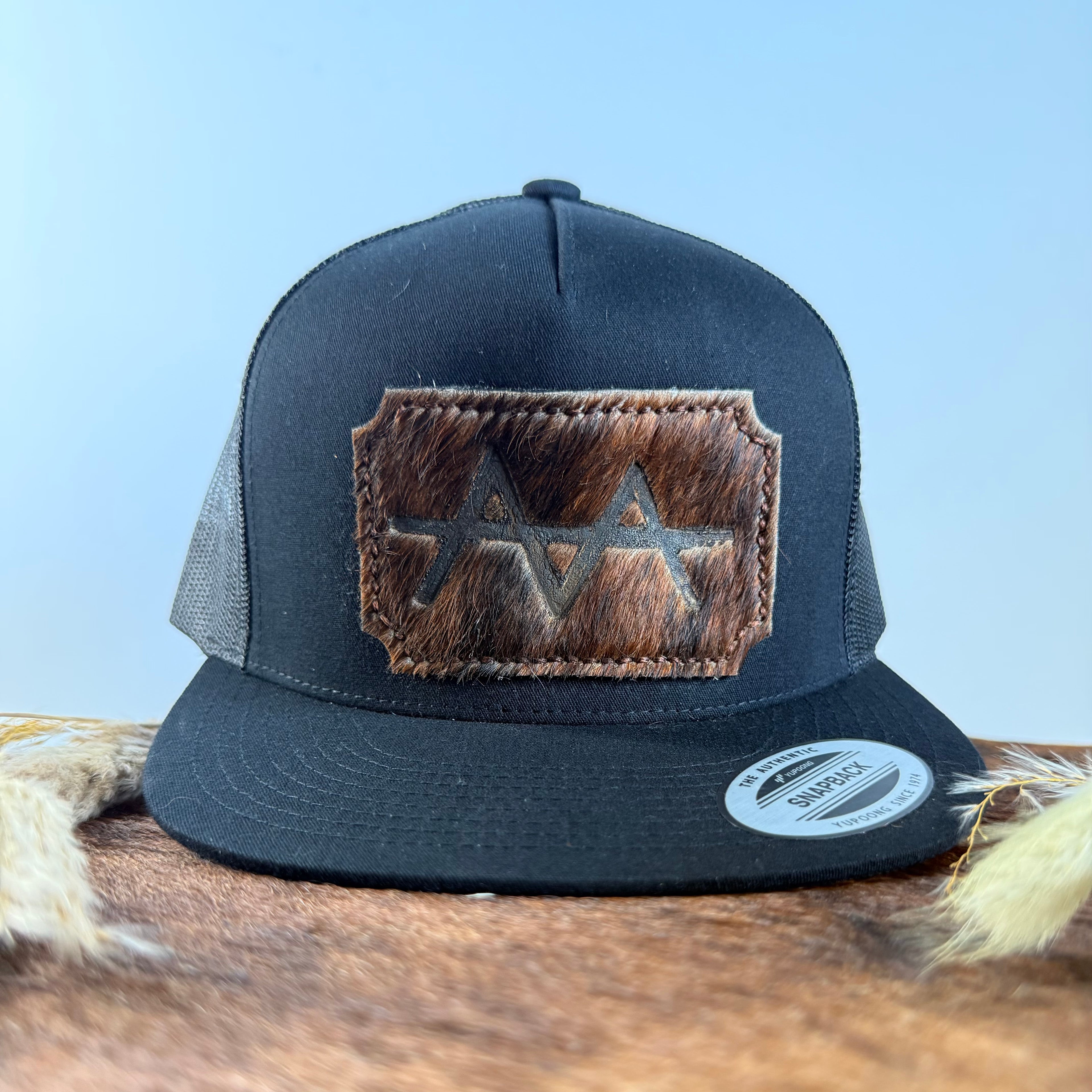 All-black trucker snapback hat with scalloped espresso brindle cowhide patch and tonal brown stitched border, branded with a registered cattle brand