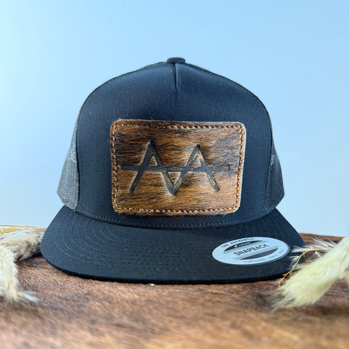 All-black trucker snapback hat with golden brown brindle cowhide patch and copper tonal stitched border, branded with a registered cattle brand