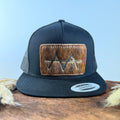 All-black trucker snapback hat with light golden tan brindle cowhide patch and red hand-stitched border, branded with a registered cattle brand