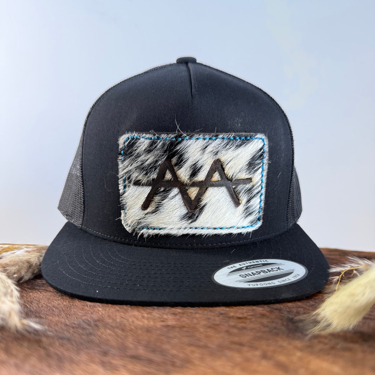 All-black trucker snapback hat with bold black and white speckled cowhide patch and electric blue hand-stitched border, branded with a registered cattle brand