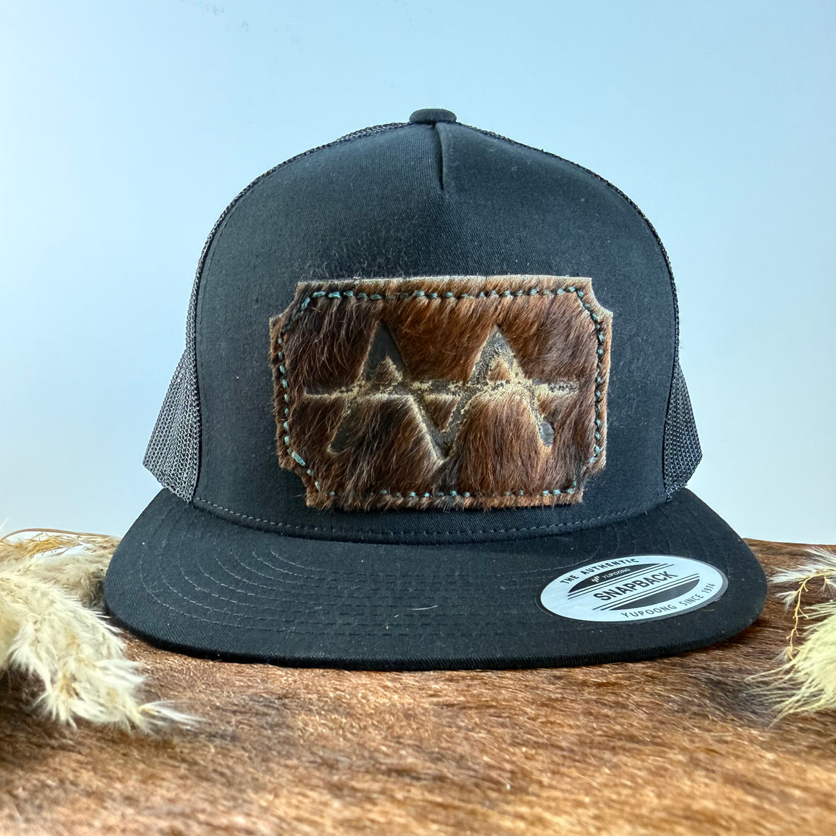 All-black trucker snapback hat with scalloped dark brindle cowhide patch and teal hand-stitched border, branded with a registered cattle brand